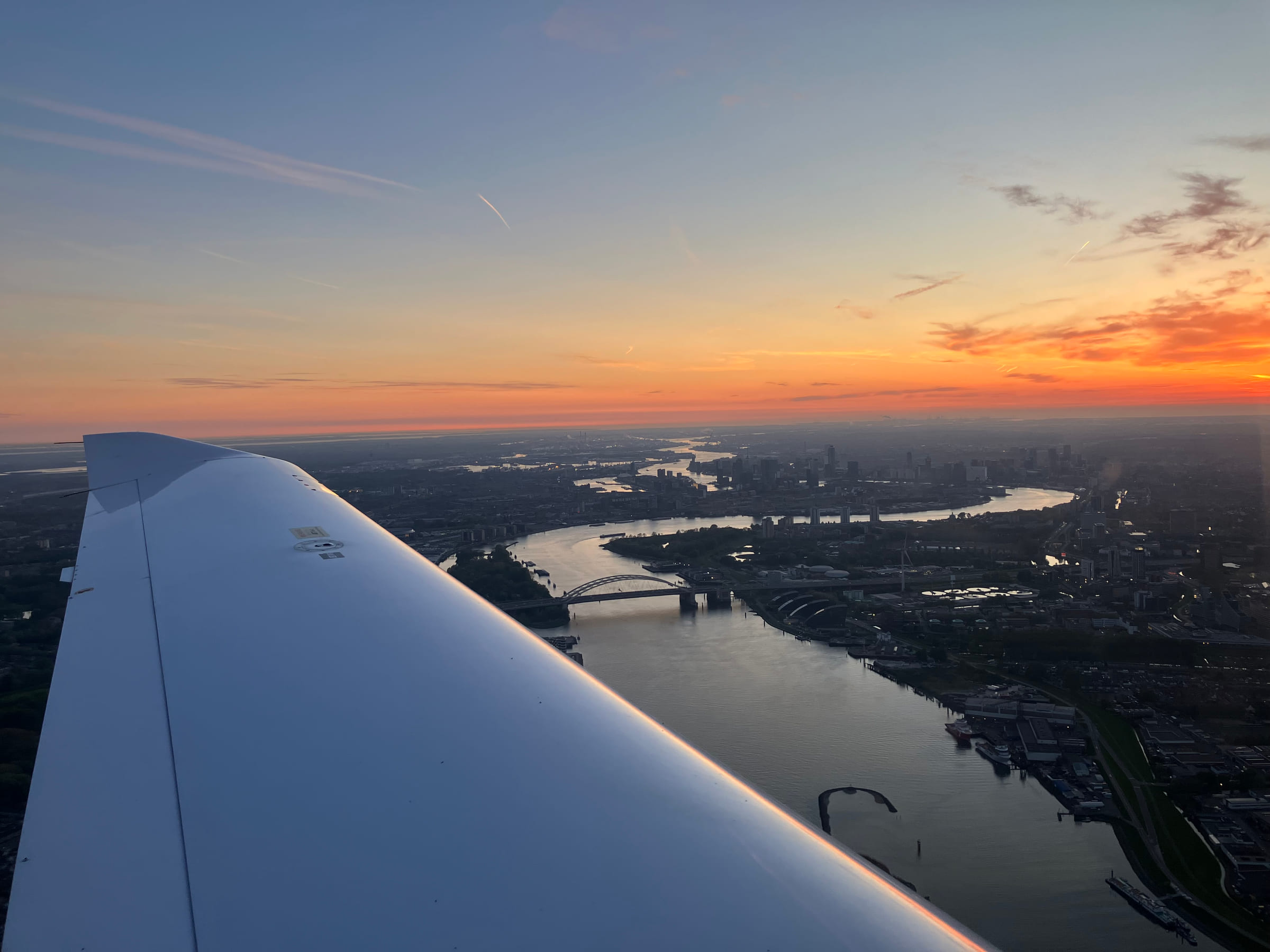 The wing of a light aircraft over the Nieuwe Maas at sunset, with the Rotterdam skyline on the horizon.